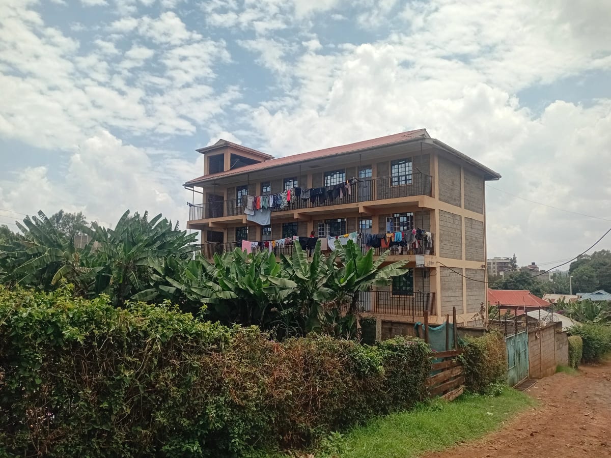 Multi-story residential building with red corrugated roof and green compound fence
