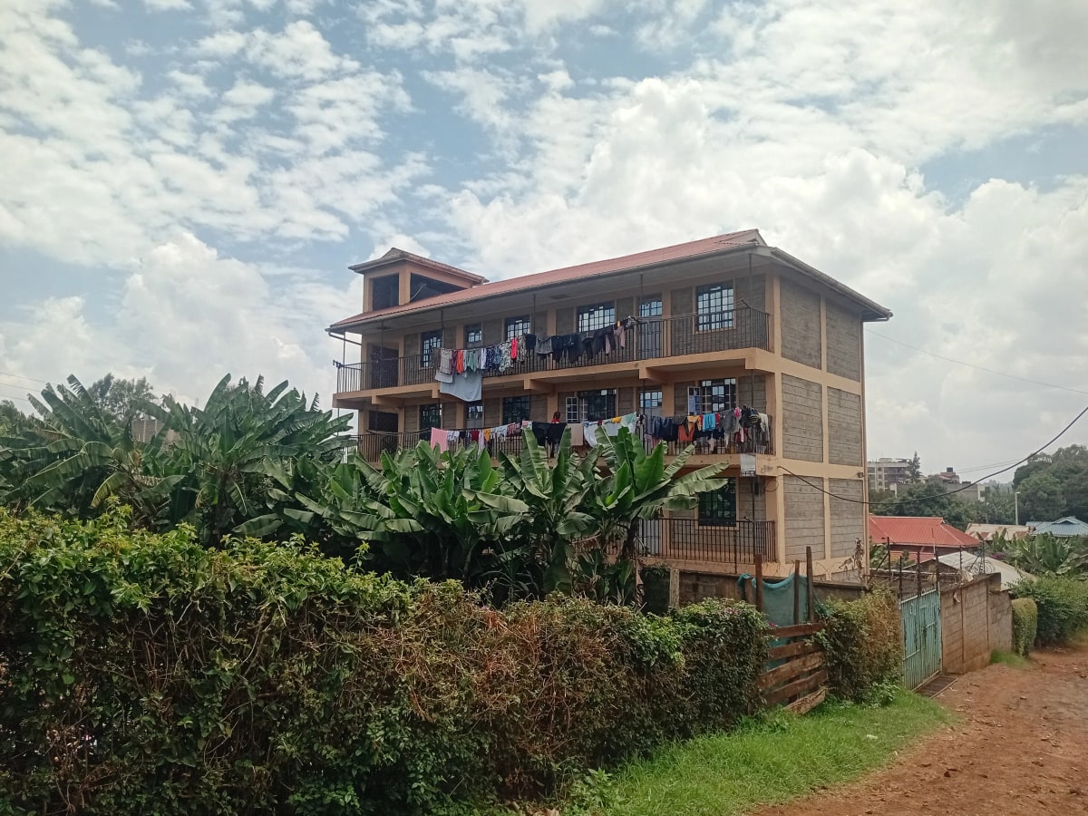 Multi-story residential building with red corrugated roof and green compound fence
